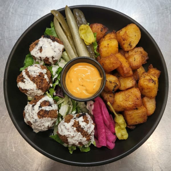 A black bowl filled with falafel, roasted potatoes, pickles, and a side of orange sauce.