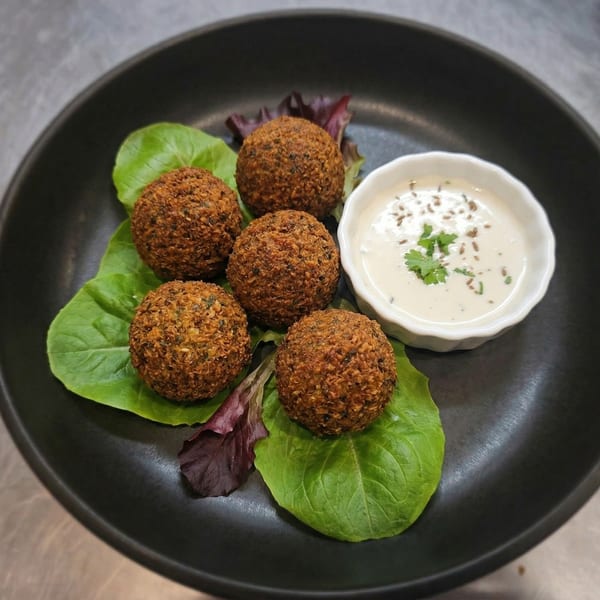 Five falafel balls on lettuce leaves with a side of white sauce, garnished with cumin seeds and cilantro.