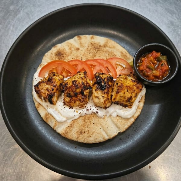 Grilled chicken skewers on pita bread with white sauce, sliced tomatoes, and a side of salsa.