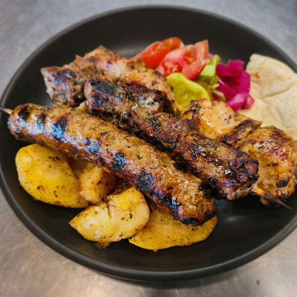Grilled meat skewers, roasted potatoes, fresh tomatoes, pickled vegetables, and pita bread on a black plate.