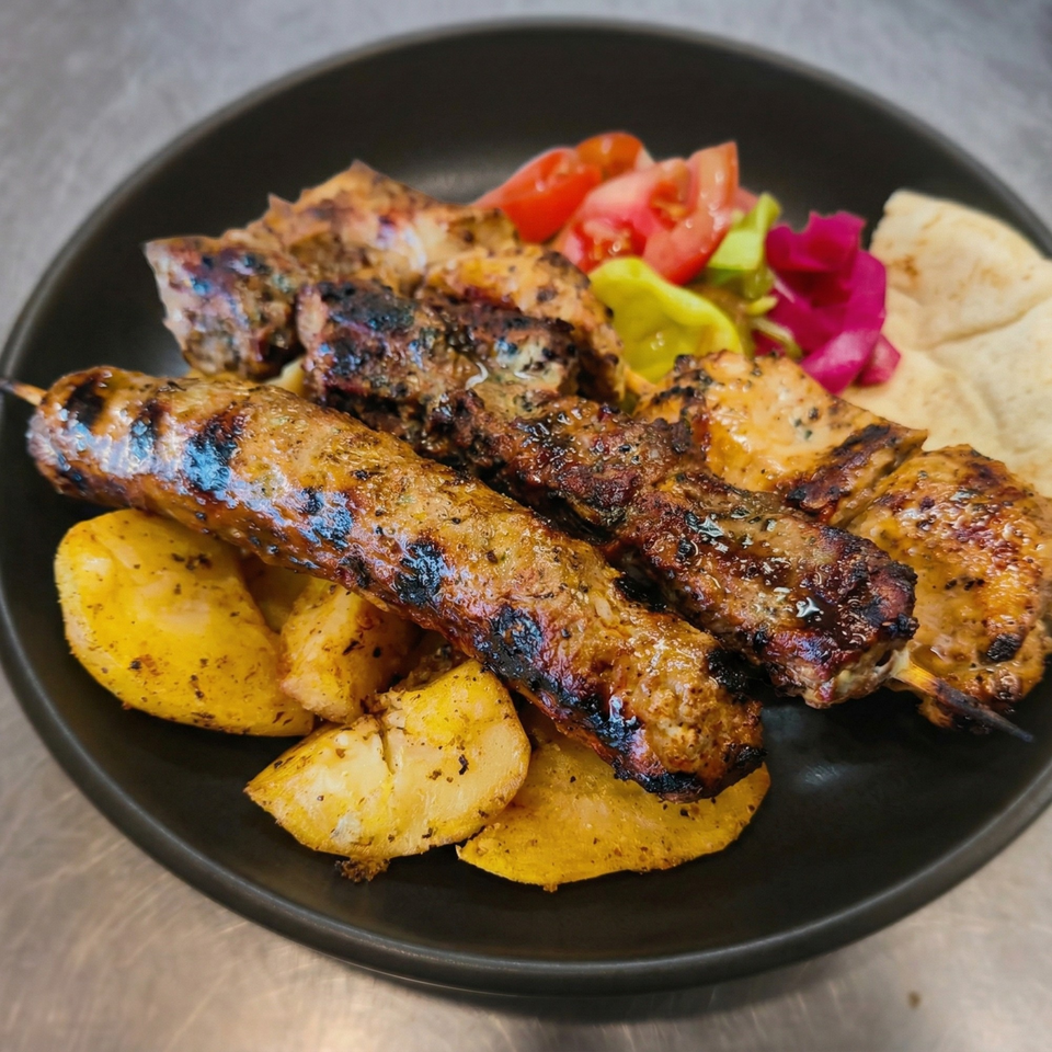 Grilled meat skewers, roasted potatoes, fresh tomatoes, pickled vegetables, and pita bread on a black plate.
