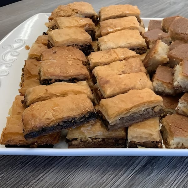 A white platter filled with rows of golden-brown baklava pastries with dark nut filling.