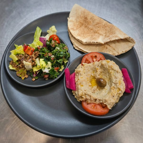 A black plate with lentil salad, baba ghanoush with tomatoes and pickles, and pita bread.